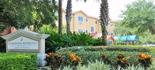 Exterior view of Homewood Residence at Freedom Plaza, a senior living community surrounded by lush greenery, palm trees, and well-maintained landscaping under a bright sky.