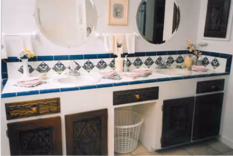 Two-sink bathroom vanity with round mirrors, tile backsplash, cabinet doors, and laundry basket.
