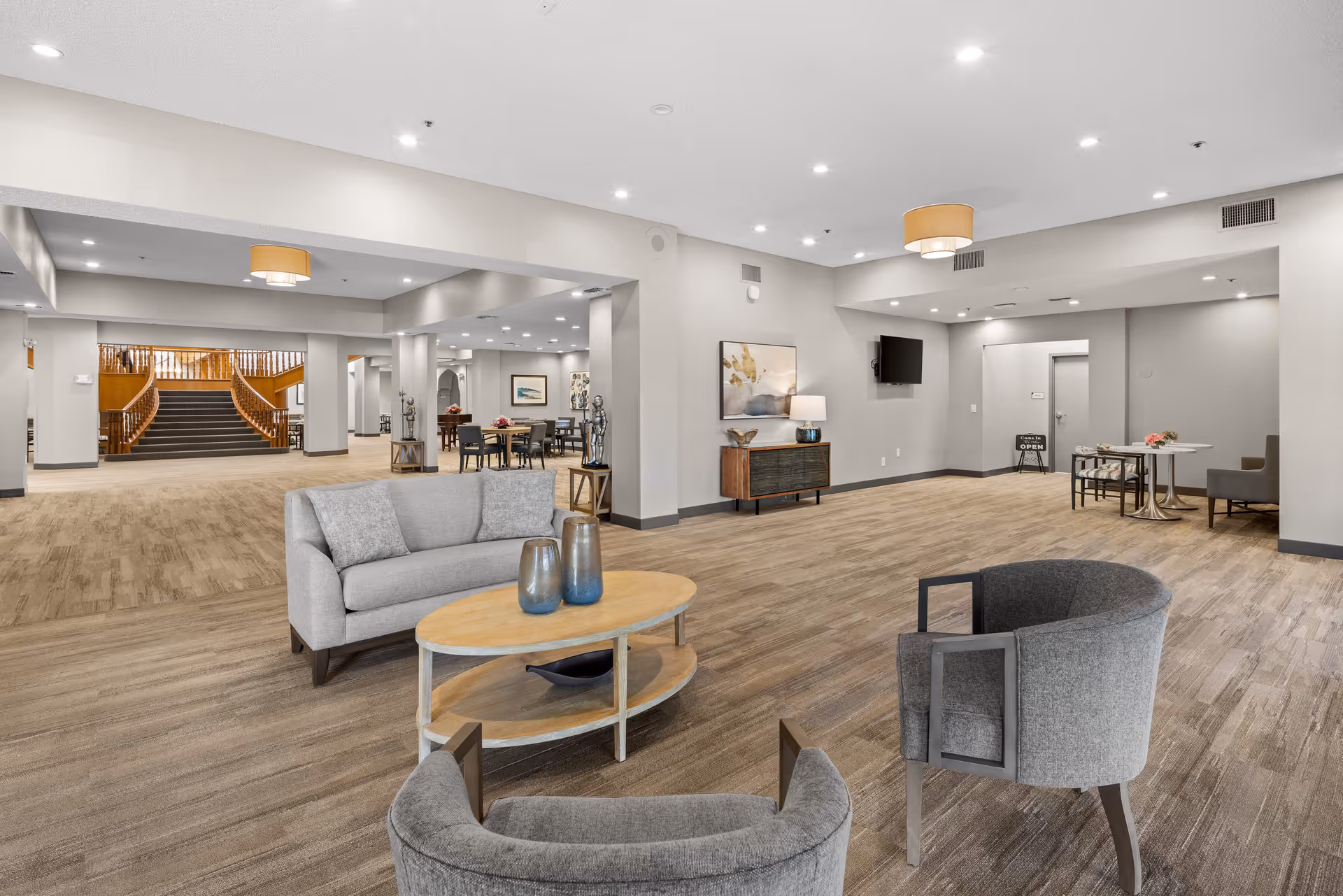 A spacious and modern senior living community common area with comfortable seating including a gray sofa and two gray armchairs around a wooden coffee table. The room features wood flooring, neutral-colored walls, recessed lighting, and decorative elements such as vases and artwork. In the background, there is a dining area with tables and chairs, a staircase with wooden railings, and a small table with a sign indicating the community is open.
