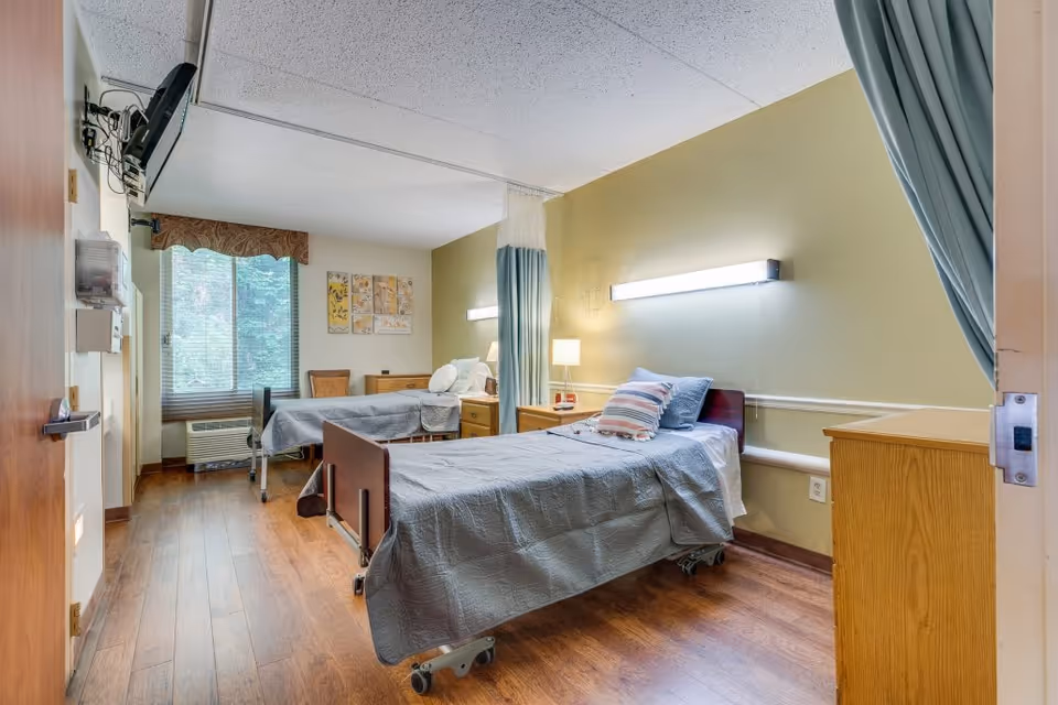 A well-lit room with two hospital-style beds, each with a pillow and blanket. The room has wooden flooring, a window with blinds and a valance, a wall-mounted TV, bedside tables with lamps, and a privacy curtain between the beds. The walls are painted light green and beige, and there is a wooden dresser near the door.