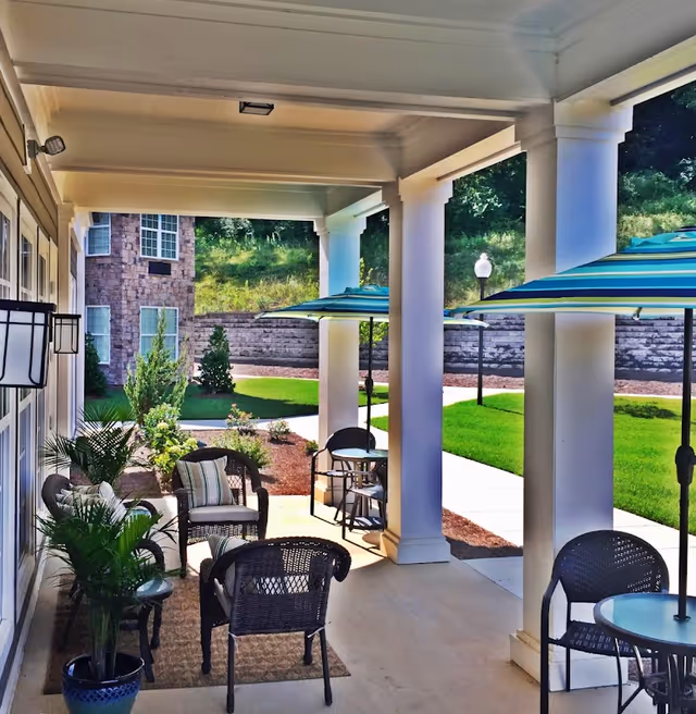 Covered outdoor patio area with wicker chairs, small tables, potted plants, and striped umbrellas. The patio overlooks a landscaped garden with green grass, shrubs, and a stone retaining wall. The building exterior features large white columns and windows.