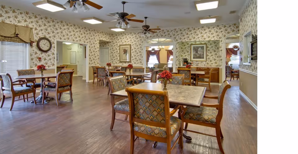 A spacious dining room in an assisted living facility with several wooden tables and cushioned chairs arranged neatly. The walls are decorated with floral wallpaper and framed artwork. Ceiling fans and lights are mounted on the ceiling. Small flower arrangements are placed on each table, and windows with blinds and valances allow natural light into the room.