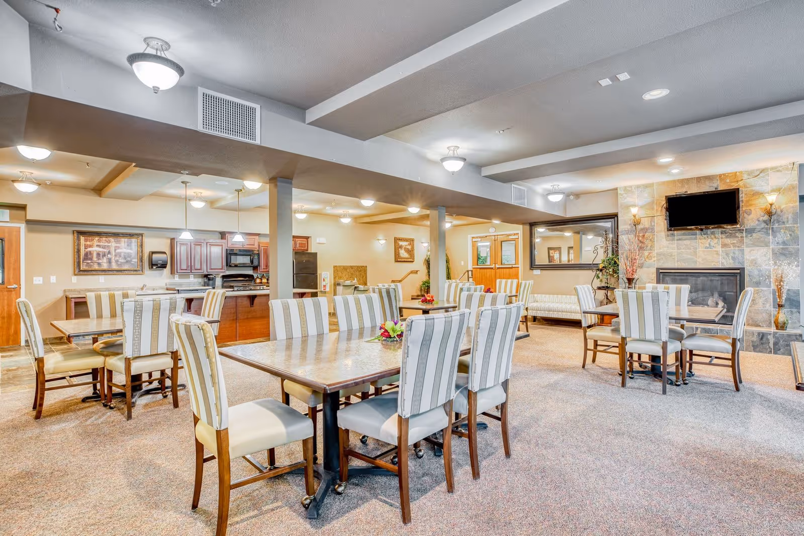 A spacious senior living dining area with multiple tables and chairs arranged neatly. The room features a fireplace with a mounted TV above it, a large mirror on the wall, and a kitchen area with wooden cabinets and appliances in the background. The space is well-lit with ceiling lights and has a warm, inviting atmosphere.