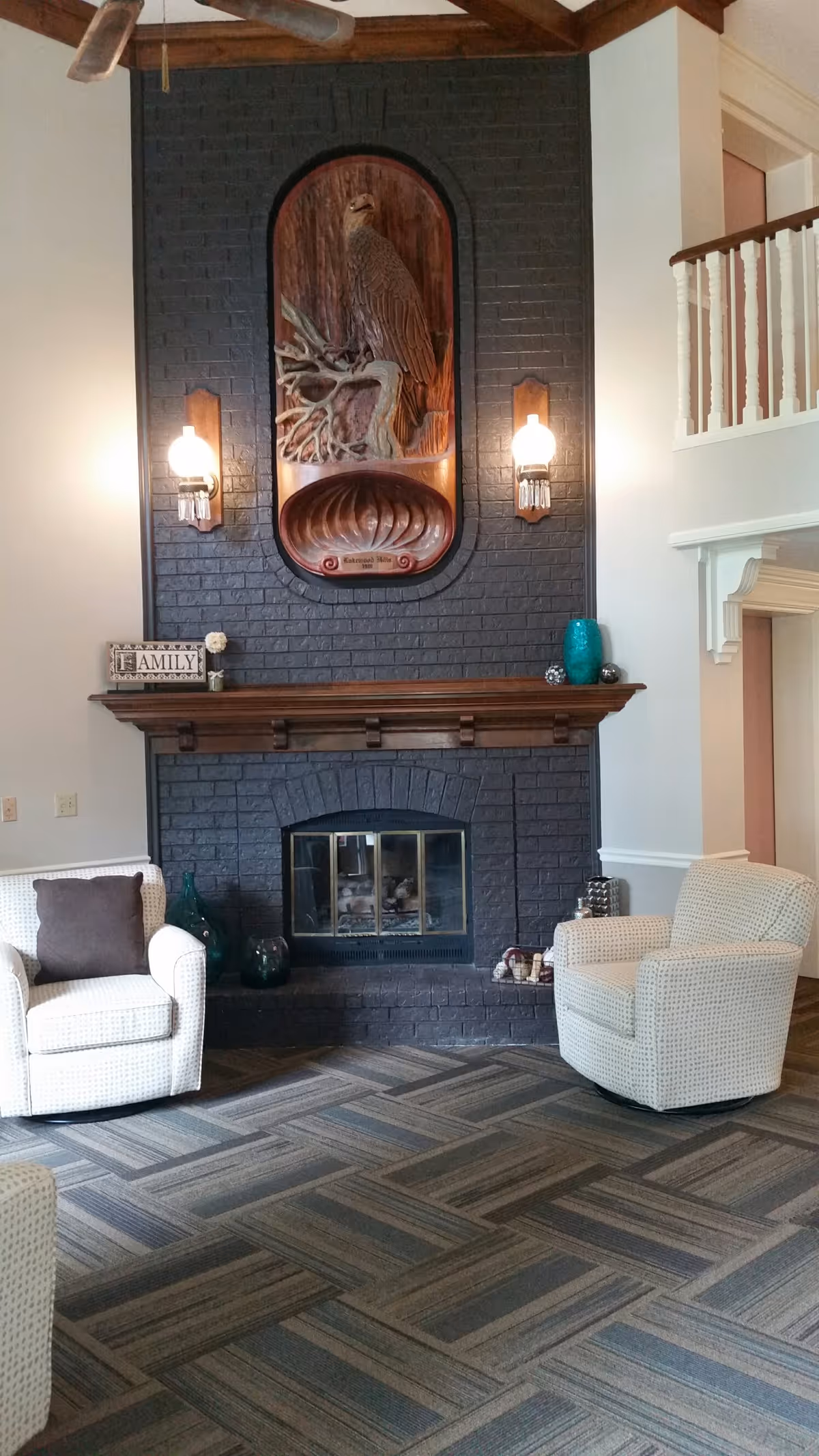 A cozy living room area with a dark brick fireplace as the focal point. Above the fireplace is a large wooden carving of a bird perched on a branch, flanked by two wall-mounted lights. On the mantel are decorative items including a sign that says 'FAMILY' and some vases. Two cushioned armchairs with patterned upholstery are positioned on either side of the fireplace on a carpeted floor with a geometric pattern. The room features a high ceiling with a ceiling fan and a white railing balcony visible on the right side.