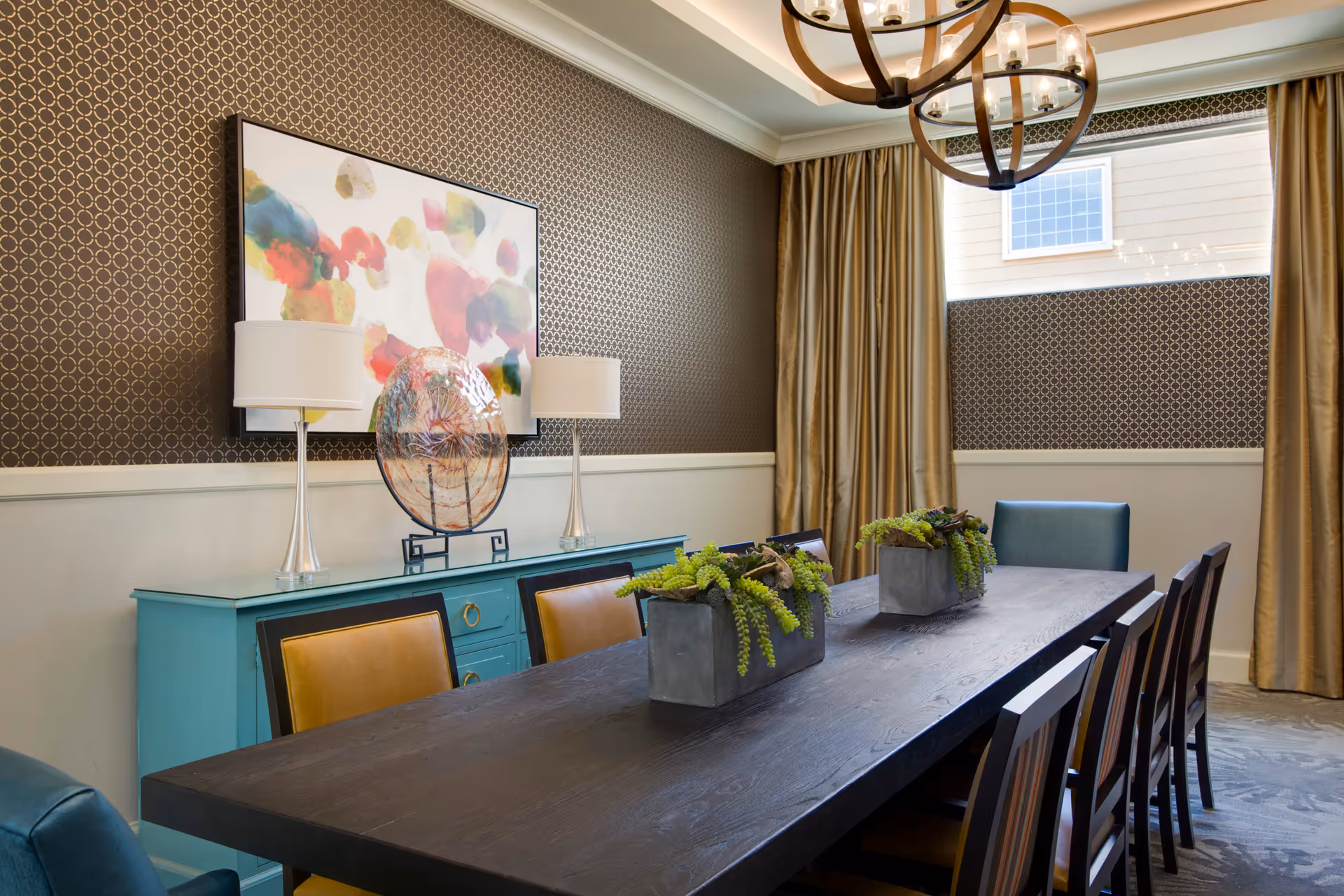 A dining room with a long dark wooden table surrounded by chairs. The table has two rectangular planters with green plants. Behind the table is a turquoise sideboard with two lamps and a decorative glass plate. Above the sideboard is a colorful abstract painting. The walls have a patterned wallpaper on the upper half and are painted white on the lower half. There is a window with gold curtains and a modern chandelier hanging from the ceiling.