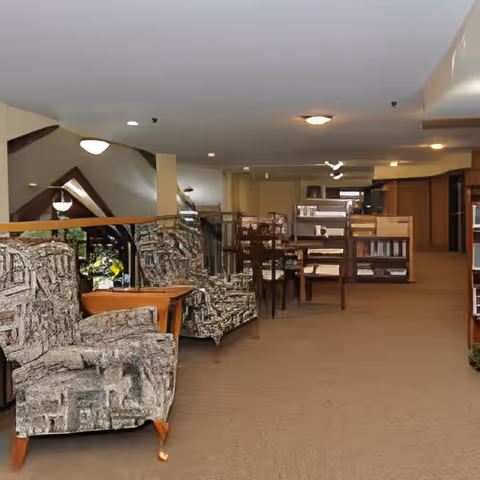 Interior view of a senior living facility lounge area with patterned armchairs, wooden tables, and bookshelves. The space is carpeted and well-lit with ceiling lights, featuring a cozy and quiet atmosphere.