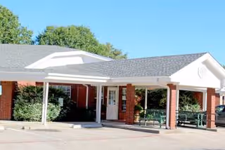 Front entrance of a brick senior living building with a covered porte-cochère, columns, benches, and landscaping.