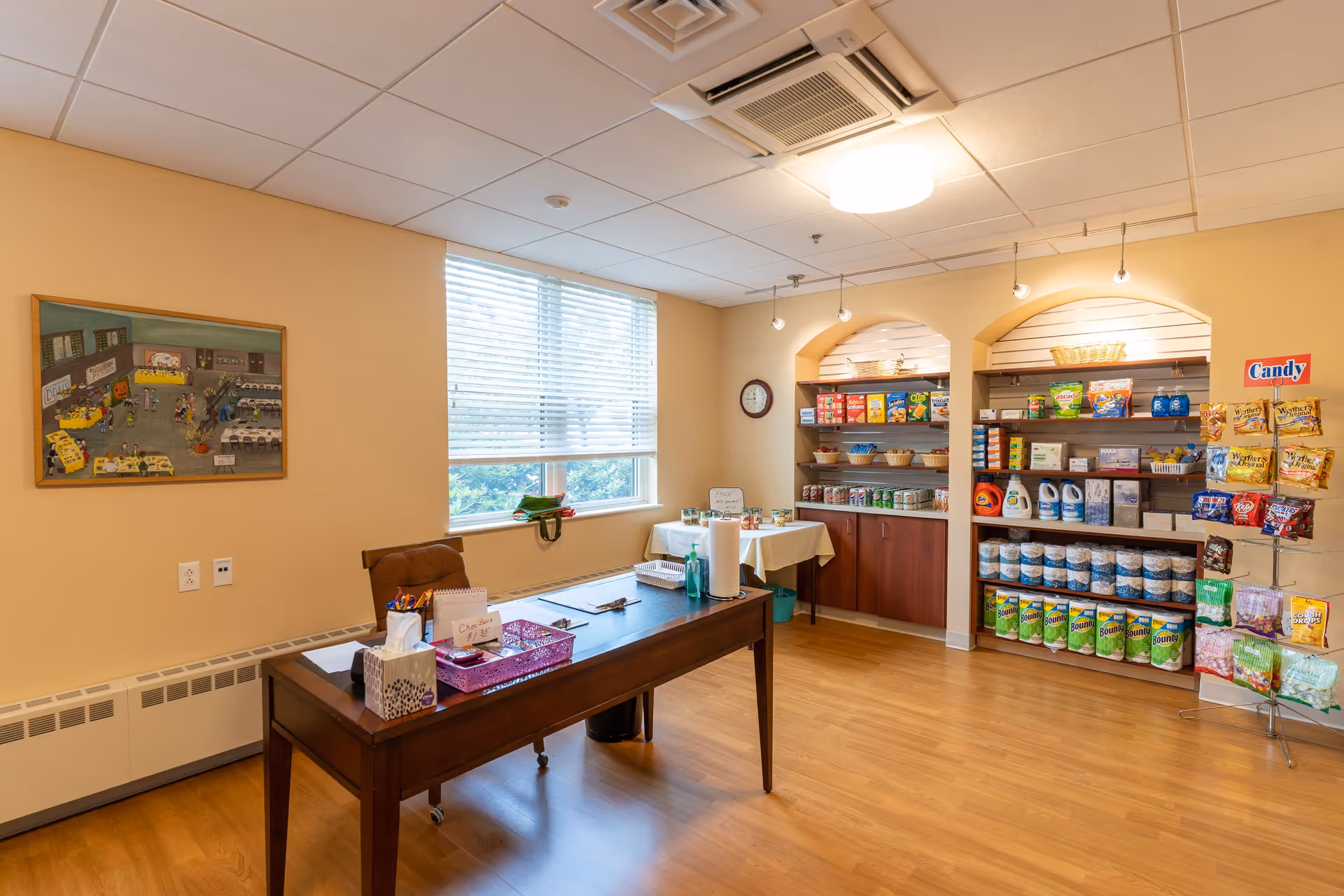 A small room with wooden flooring and beige walls featuring a wooden desk with office supplies and a tissue box. Behind the desk, there is a window with blinds partially open, letting in natural light. On the right side, there are shelves stocked with various grocery items including snacks, canned goods, paper towels, and cleaning supplies. A small table with a white tablecloth holds additional items. A clock is mounted on the wall above the shelves, and a colorful painting hangs on the left wall.