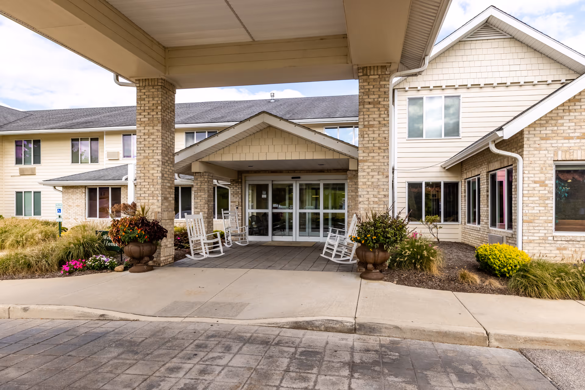 Covered front entrance of a senior living facility with rocking chairs, large planters, and glass double doors under a porte-cochere.