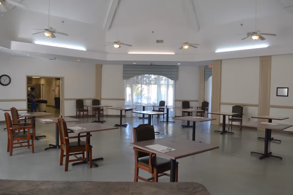 A spacious dining room with multiple square tables and chairs arranged with social distancing. The room has a high ceiling with ceiling fans and large windows with sheer curtains allowing natural light to enter. A clock is visible on the wall near an open doorway leading to another room.