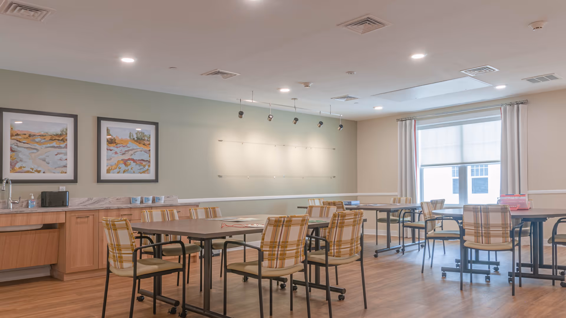 A well-lit room with multiple tables and chairs arranged for group seating. The chairs have a plaid pattern with yellow and beige tones. The room features wooden flooring, light-colored walls, two framed landscape paintings on one wall, and a large window with white curtains and a roller shade. There is a countertop with a sink and some items on it along one wall.