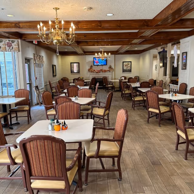 A dining room with multiple square tables and cushioned chairs arranged neatly. The room features wooden ceiling beams, two chandeliers, a fireplace with fall decorations, framed pictures on the walls, and a television mounted above the fireplace. The floor is tiled with a wood-like pattern, and windows with floral valances allow natural light into the space.