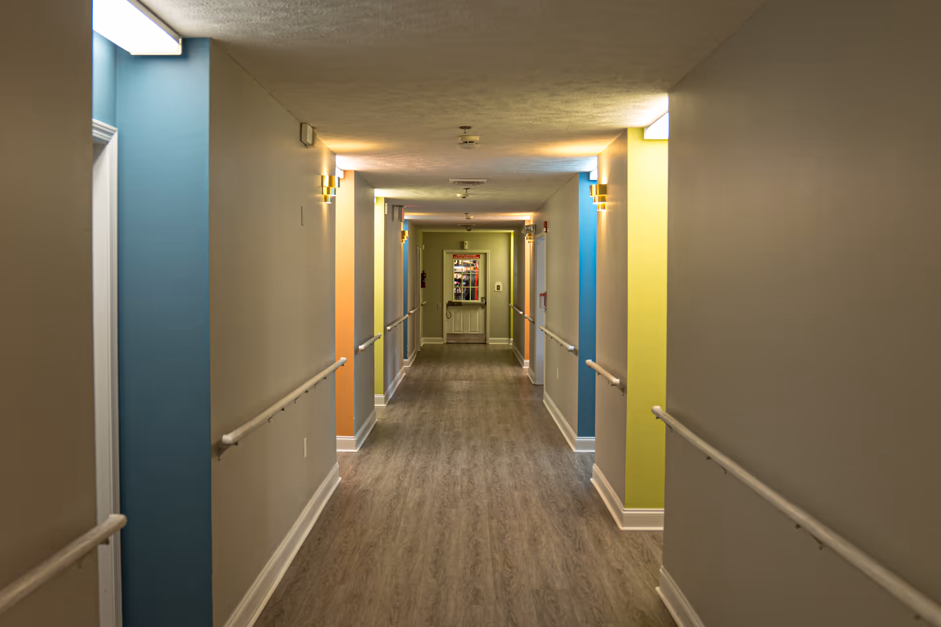 A long indoor hallway in an assisted living facility with handrails on both sides, colorful wall sections in blue, orange, and yellow, and overhead lighting. The floor is wood-style laminate and there is a door at the end of the hallway.