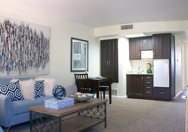 A modern living area in a senior living facility featuring a blue sofa with patterned and fluffy pillows, a wooden coffee table with decorative items, a small dining table with two chairs, and a kitchenette with dark wood cabinets, a small refrigerator, and a sink. The walls are light-colored with framed artwork, and the floor is carpeted.