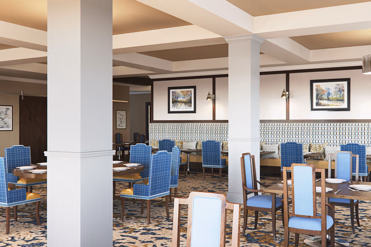 A spacious dining room with multiple round and rectangular wooden tables set with white plates and silverware. The chairs have blue upholstery with a checkered pattern and wooden frames. The room features white columns, patterned carpet, and framed landscape paintings on the walls. There is a cushioned bench along one wall with patterned pillows.