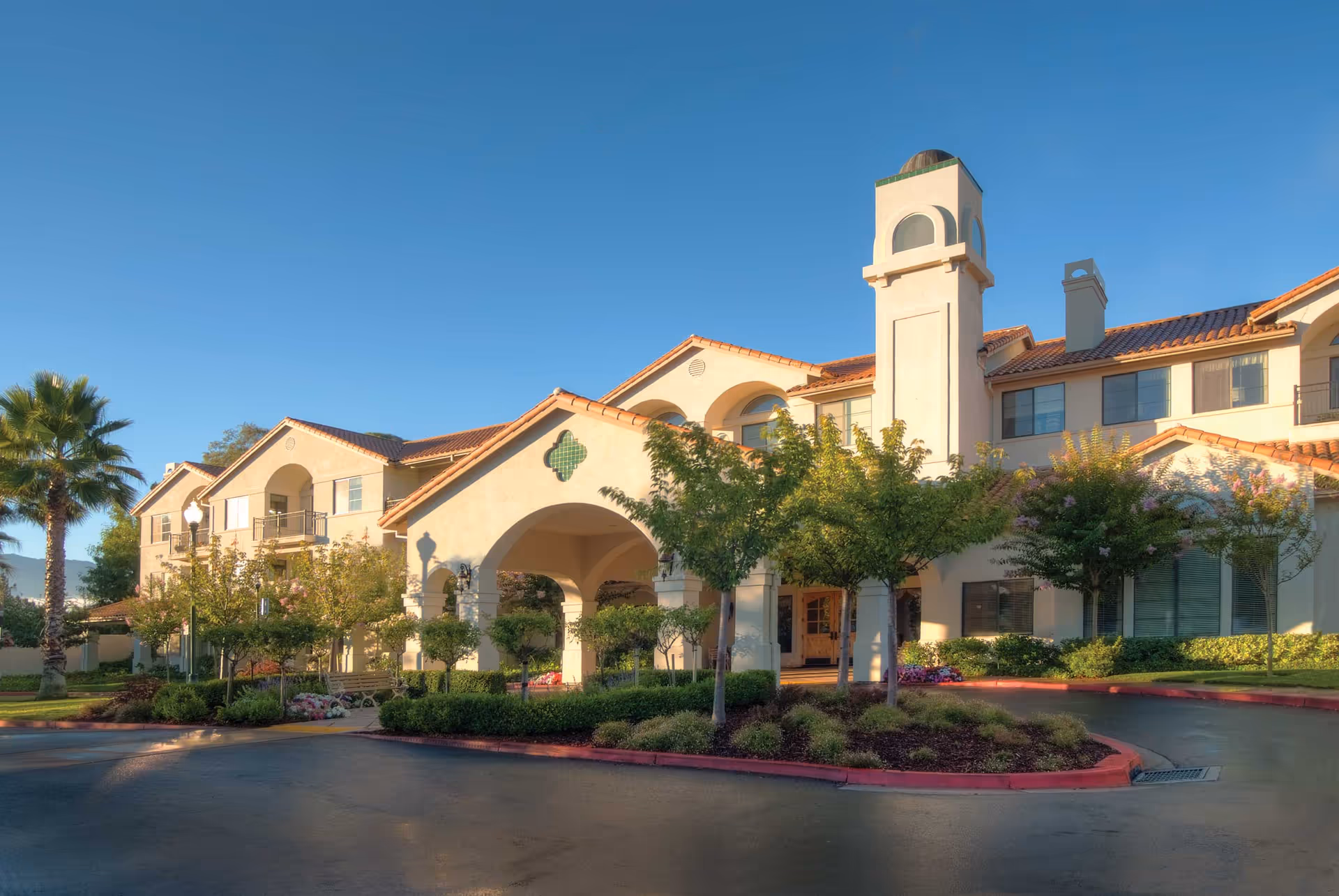 Exterior view of Oakmont of Montecito, a senior living facility with a Mediterranean-style building featuring a tiled roof, arched entrance, and a tower. The building is surrounded by landscaped greenery, trees, and a driveway under a clear blue sky.