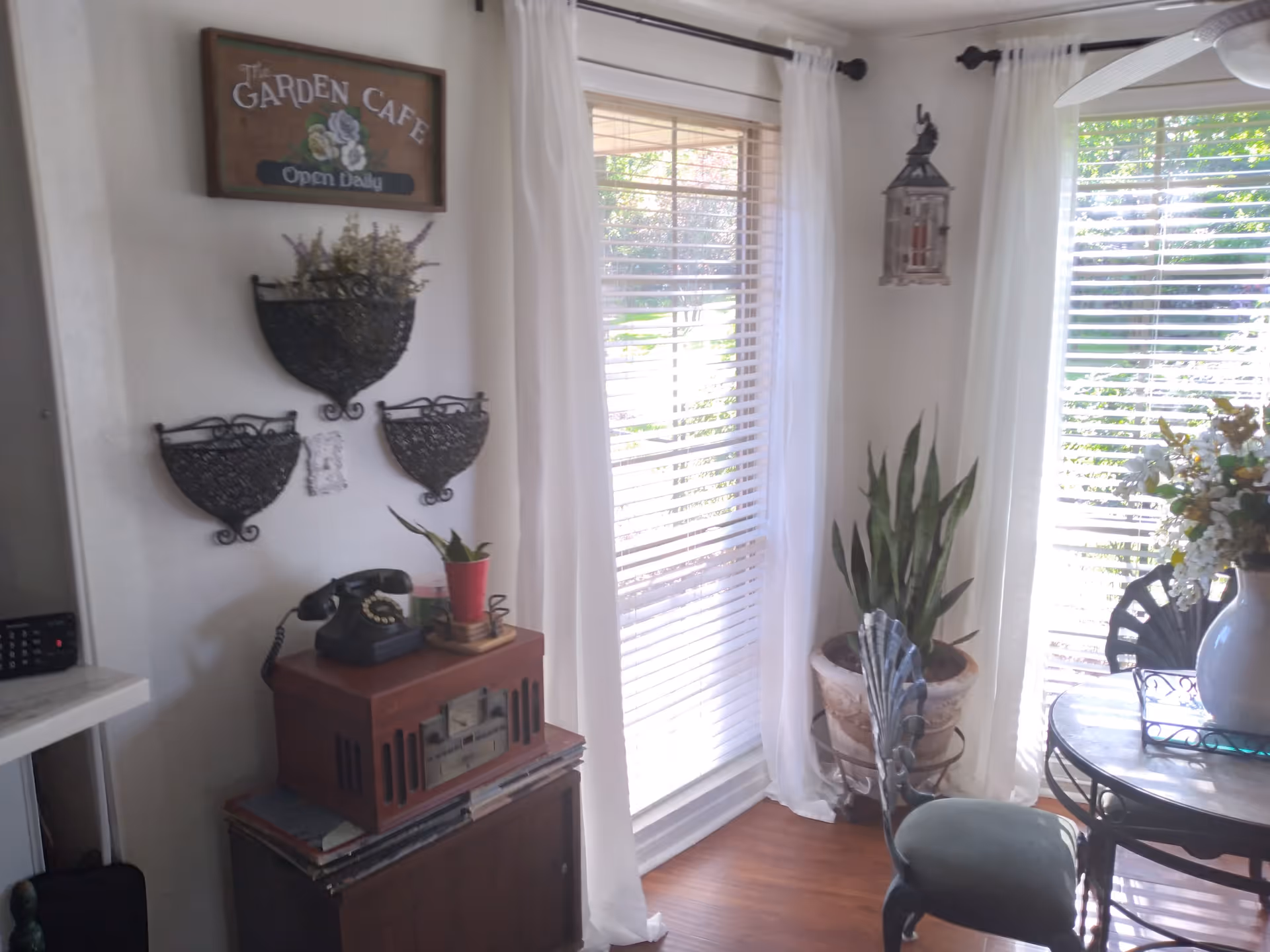 A cozy corner of a room with two large windows covered by white blinds and sheer white curtains. There is a round table with a vase of flowers and a decorative chair with a green cushion. A large potted plant is placed near the windows. On the wall, there is a wooden sign that reads 'The Garden Cafe Open Daily' above three decorative black metal wall planters. Below the planters, there is a vintage-style wooden cabinet with an old-fashioned rotary telephone and a small potted plant on top.