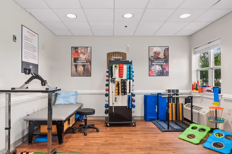 A physical therapy or rehabilitation room with exercise equipment including a padded treatment table with a pillow, a stool, a rack holding various exercise bands and weights, and other therapy tools. The room has wood flooring, white walls with two motivational posters, and a window letting in natural light.
