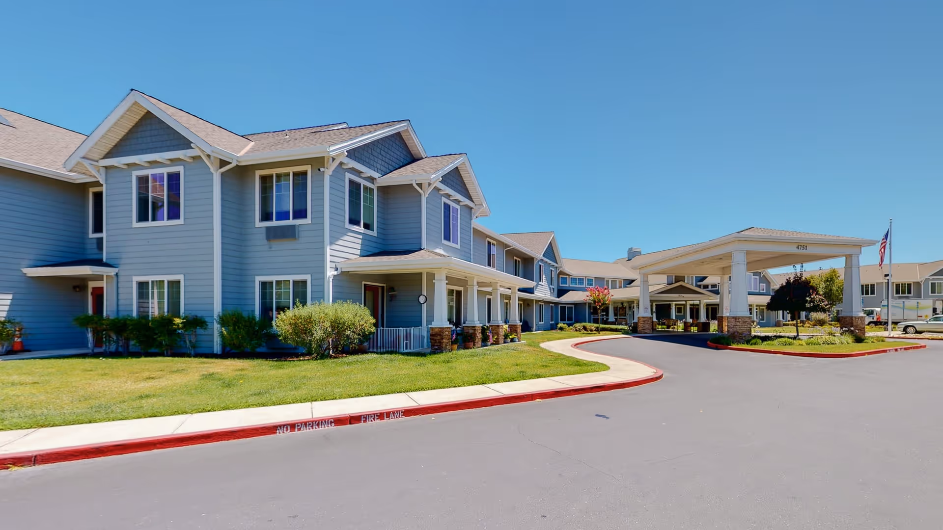 Exterior view of The Commons At Dallas Ranch senior living facility showing a two-story building with gray siding, multiple windows, a covered entrance with pillars, a driveway, and a well-maintained lawn under a clear blue sky.