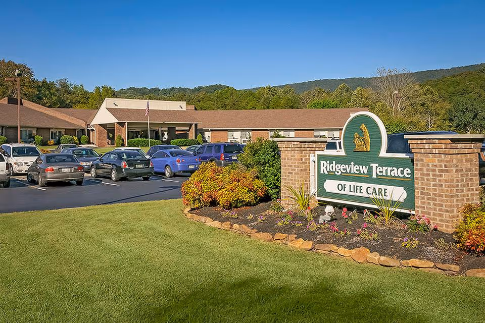 Exterior view of Ridgeview Terrace of Life Care facility with a parking lot filled with cars, a well-maintained lawn, landscaped flower beds, and a sign displaying the facility's name in front. The building has a brown roof and brick walls, with trees and hills in the background under a clear blue sky.