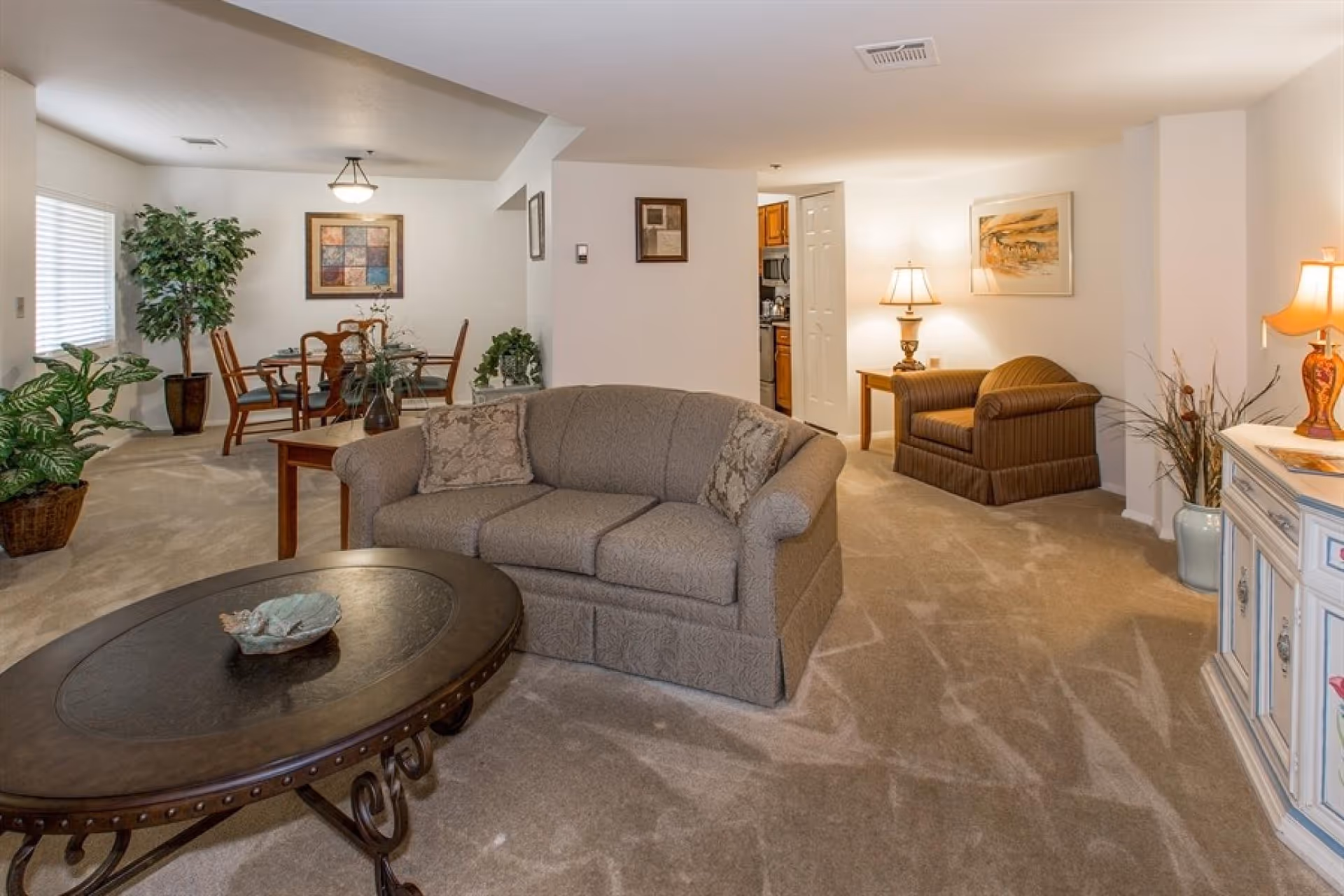 A cozy living room area with a beige patterned sofa and matching armchair, a dark wooden oval coffee table with a decorative bowl, beige carpet, and soft lighting from table lamps. In the background, there is a dining area with a wooden dining table and chairs, a large potted plant, and framed artwork on the walls. A small kitchen area is partially visible through a doorway.