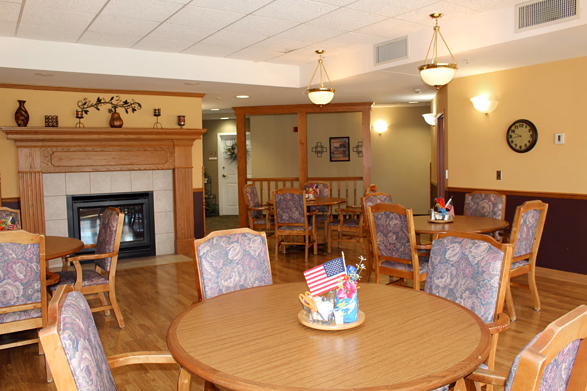 A cozy dining area in a nursing home with round wooden tables and floral upholstered chairs. Each table has a centerpiece with small American flags and decorative items. The room features a fireplace with a wooden mantle decorated with vases and candle holders. The walls are painted in warm tones with wall sconces and a clock. The floor is wooden, and there are ceiling lights providing illumination.