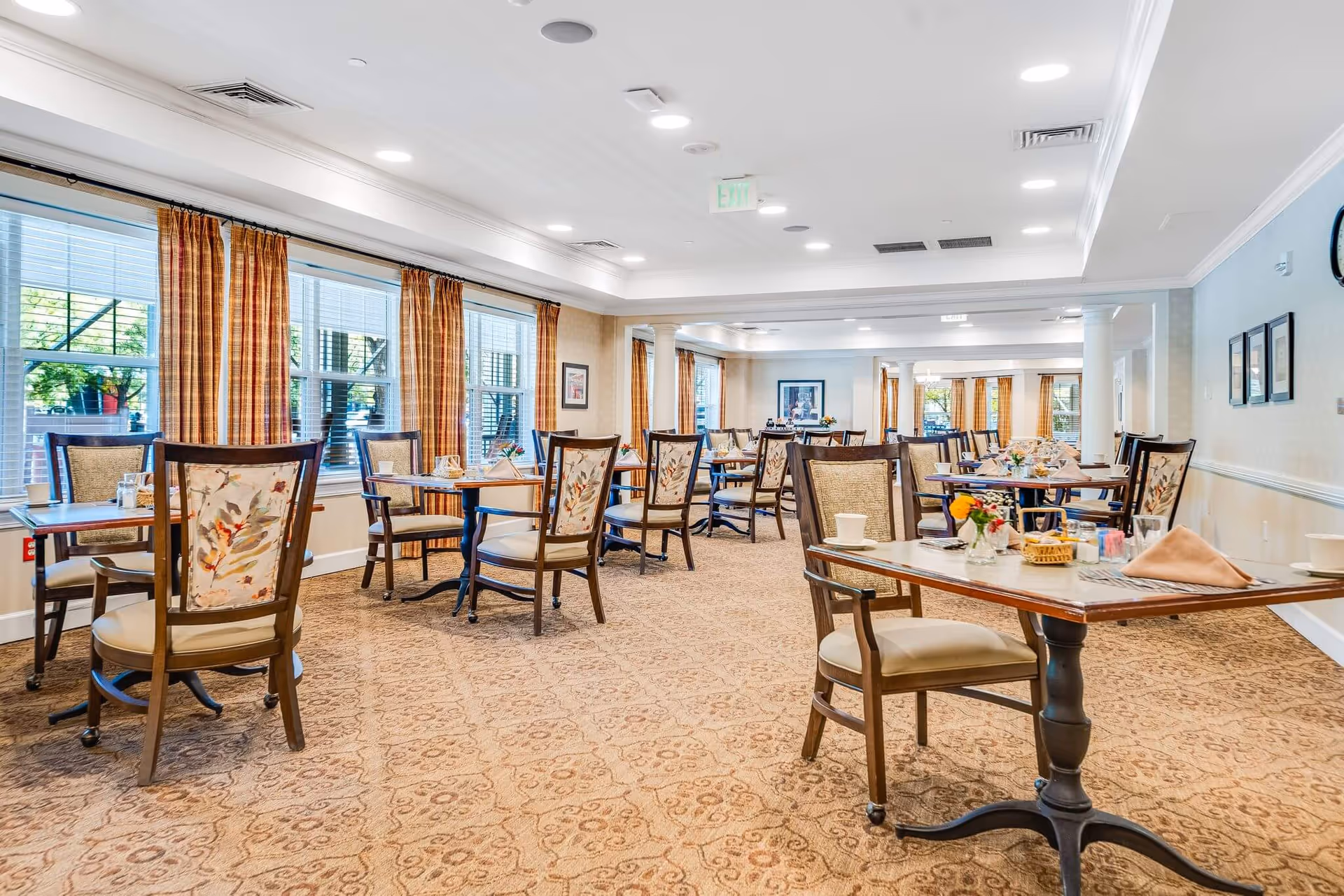 A spacious dining room with multiple tables and chairs arranged neatly. Each table is set with cups, napkins, and small flower arrangements. Large windows with orange and beige plaid curtains allow natural light to fill the room. The carpet has a patterned design, and the walls are light-colored with framed pictures. The ceiling has recessed lighting and ventilation vents.