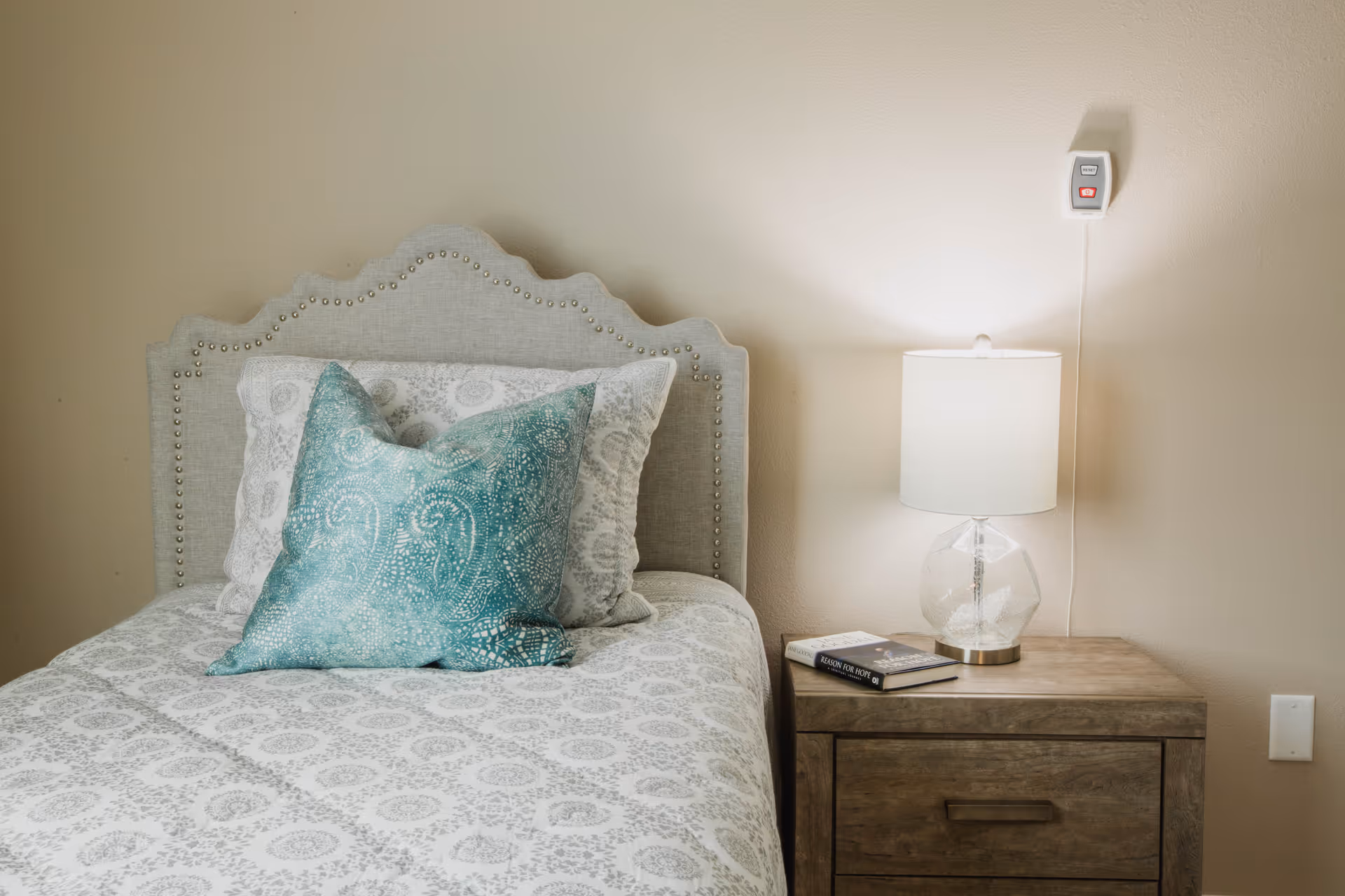 A neatly made bed with a decorative headboard and patterned bedding, featuring a teal accent pillow. Next to the bed is a wooden nightstand with a clear glass lamp and two books on top. A wall-mounted emergency call button is visible above the nightstand.