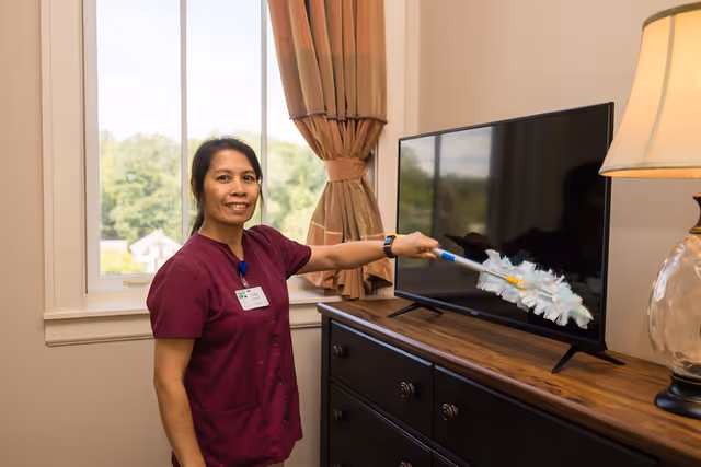 A woman wearing a maroon uniform dusts a flat-screen TV with a feather duster in a room with a window, beige walls, and a wooden dresser. There is a lamp with a beige shade on the dresser and brown curtains on the window.