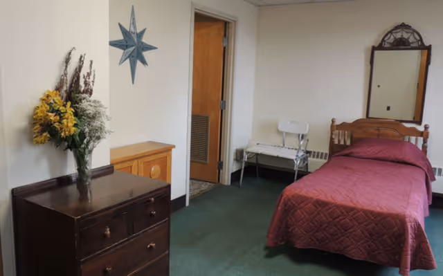 Small furnished bedroom with a single bed covered in a maroon quilt, a wooden dresser with a vase of flowers, a mirror, and a chair.