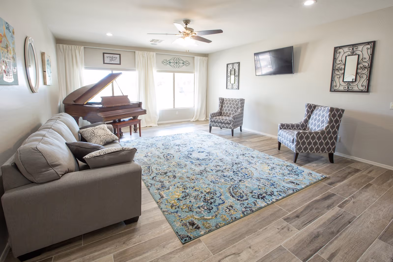 A bright living room with a gray sofa, two patterned armchairs, a large blue and beige area rug, a grand piano near two large windows with white curtains, a ceiling fan with lights, and a wall-mounted flat screen TV.