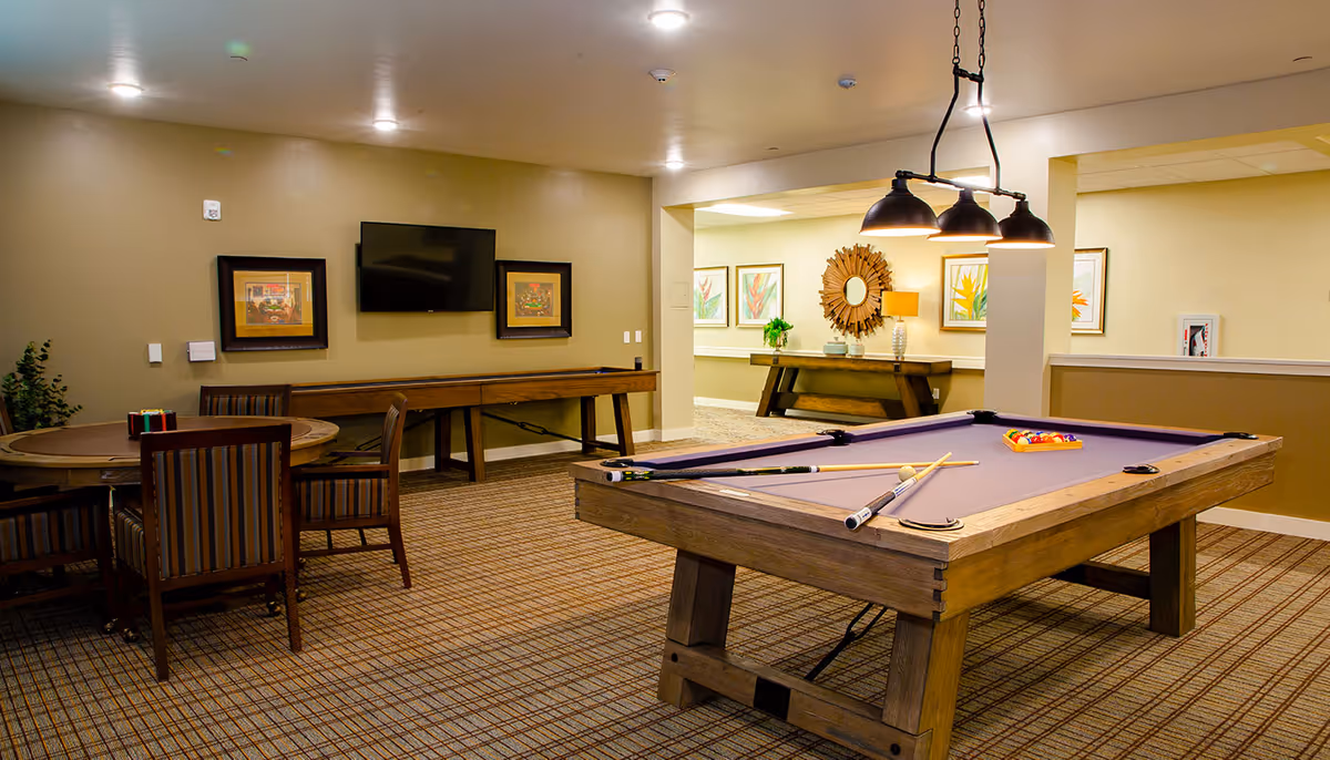 A well-lit recreational room featuring a pool table with cues and balls arranged on it, a round table with four striped chairs, a shuffleboard table against the wall, a wall-mounted flat screen TV, framed artwork, and a decorative console table with a mirror and lamp in the background.