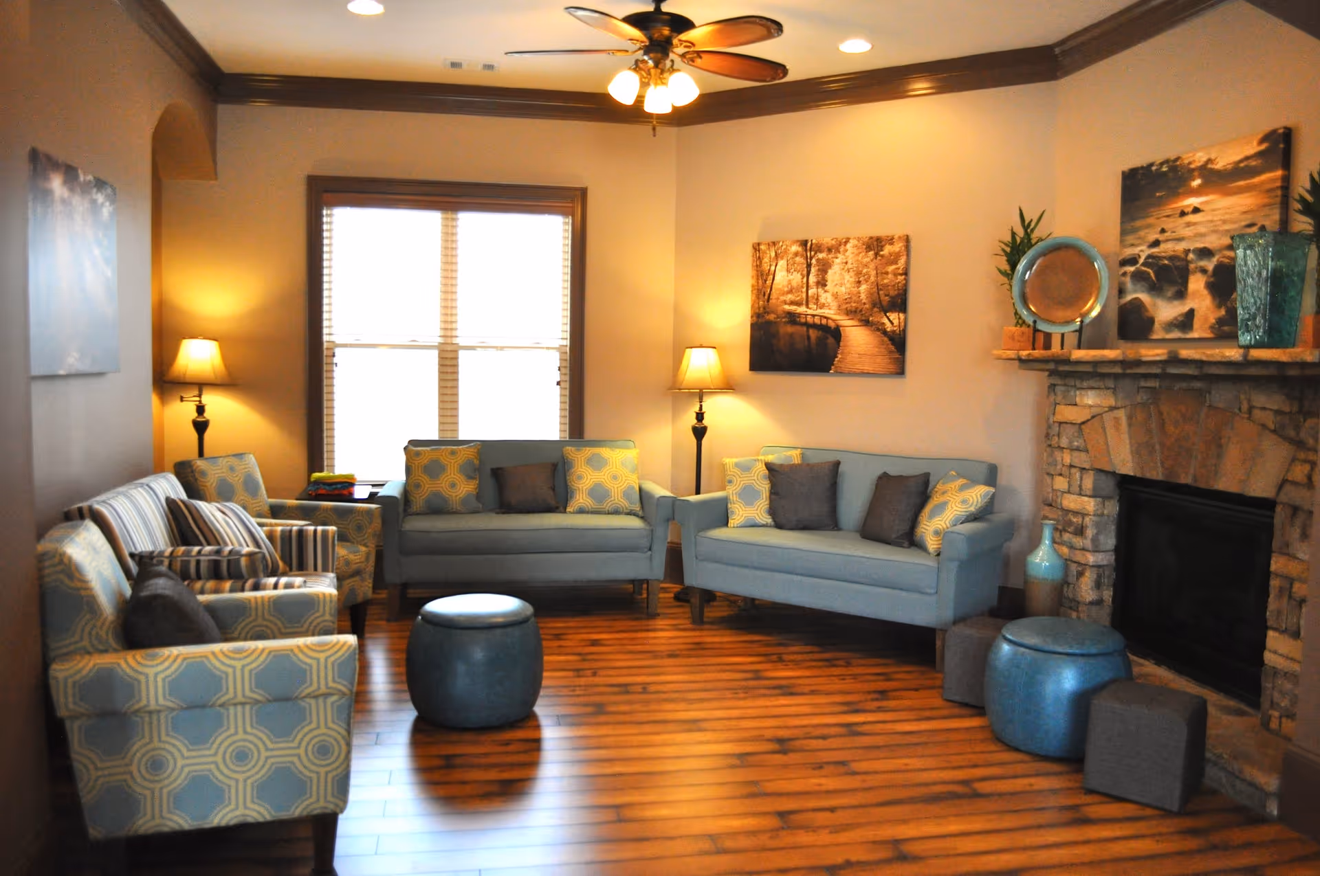 A cozy living room with wooden flooring, two blue sofas with patterned and solid cushions, two matching armchairs, a round blue ottoman, and a stone fireplace. The room is warmly lit by two floor lamps and a ceiling fan with lights. There are framed pictures on the walls and decorative plants on the fireplace mantel.