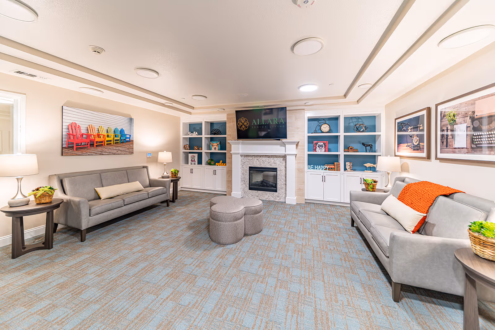 A bright and cozy senior living common area with two gray sofas facing each other, a fireplace with a TV above displaying the Allara Senior Living logo, built-in white shelves with decorative items, colorful artwork on the walls, and soft lighting from table lamps.