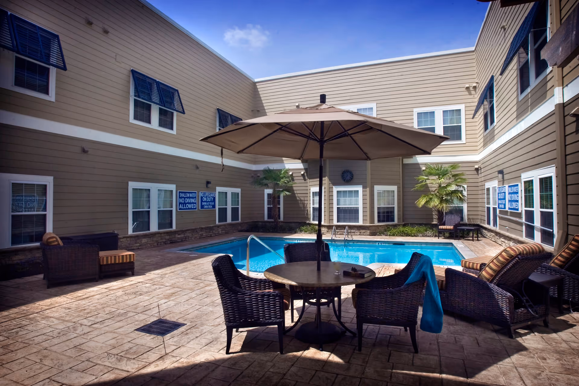 Outdoor courtyard area with a swimming pool surrounded by a two-story building. There are several wicker chairs and loungers with cushions around a round table shaded by a large umbrella. Palm plants are placed near the building walls, and signs on the windows indicate pool rules.