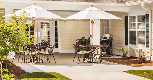 Outdoor patio at a senior living facility with tables, chairs, umbrellas and a barbecue by the building entrance.