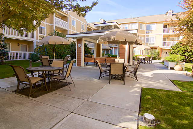 Outdoor courtyard area at Brookdale Richland featuring multiple tables with umbrellas and chairs on a concrete patio, surrounded by a multi-story residential building with balconies and greenery.