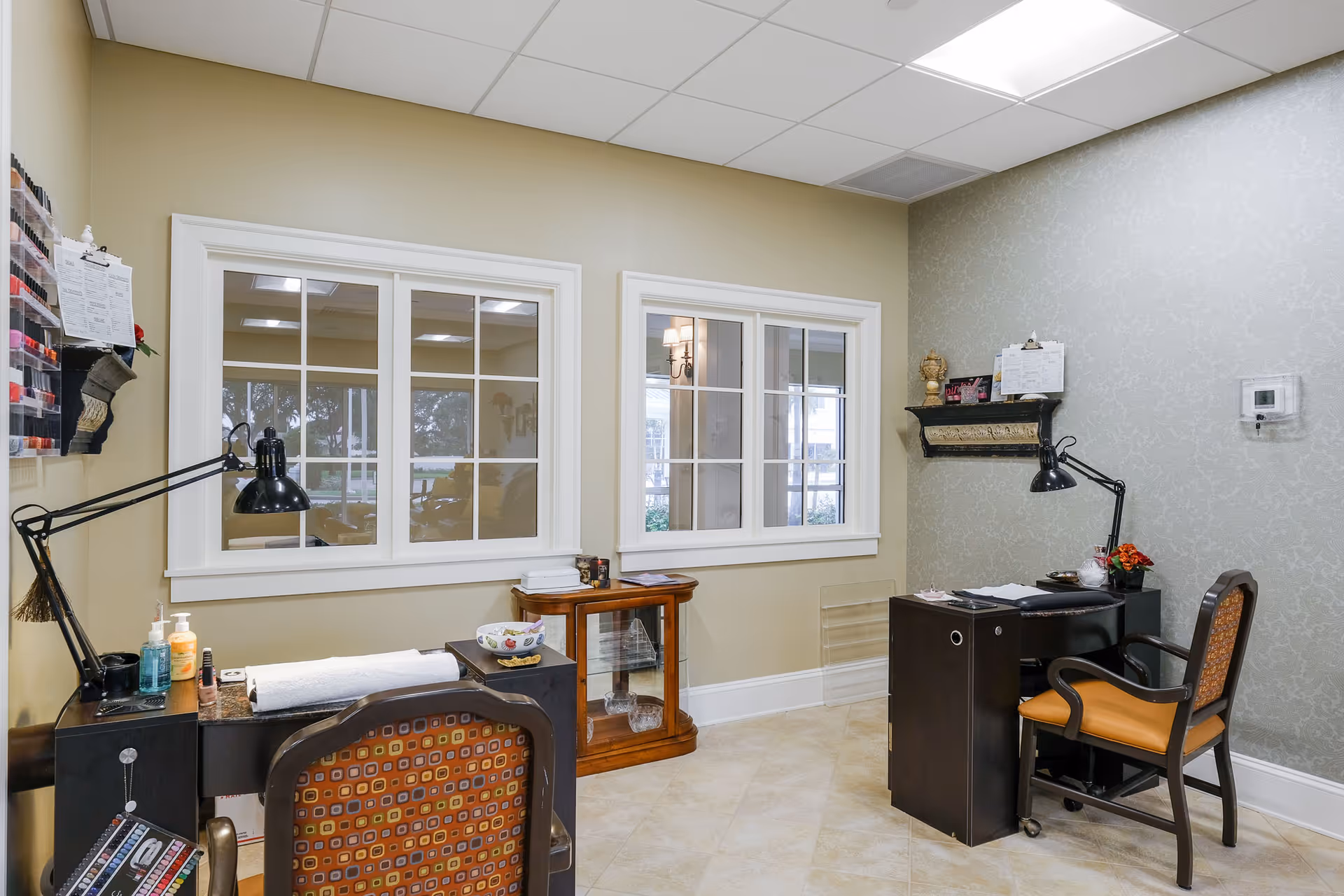 Interior view of a nail salon area with two manicure stations, each equipped with a black adjustable desk lamp, chairs with patterned upholstery, and nail polish displays on the wall. The room has beige walls, tiled floor, and two windows with white frames looking into another room.