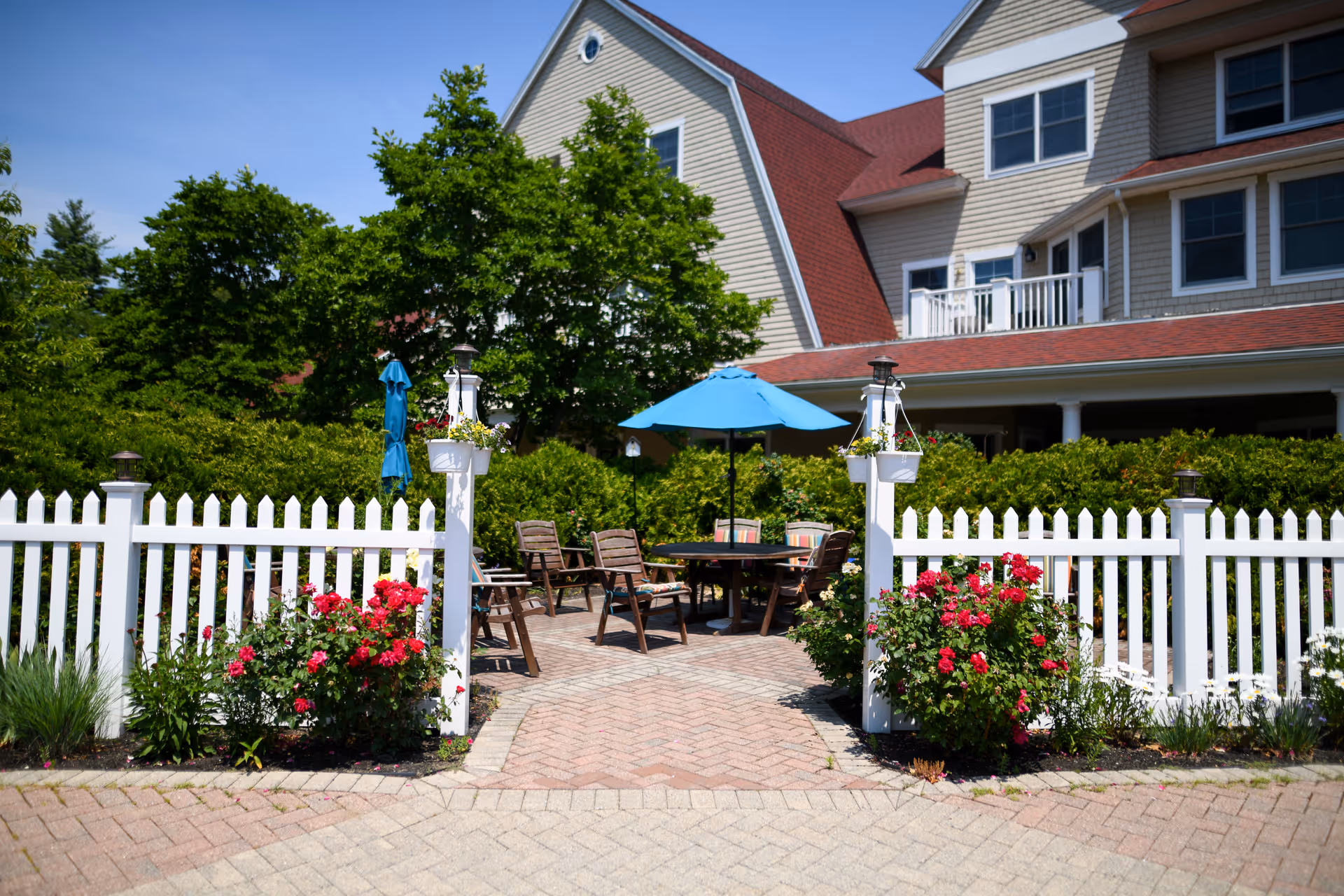 Outdoor seating area with wooden chairs and a table under a blue umbrella, surrounded by a white picket fence and blooming red flowers, with a large building and green trees in the background under a clear blue sky.