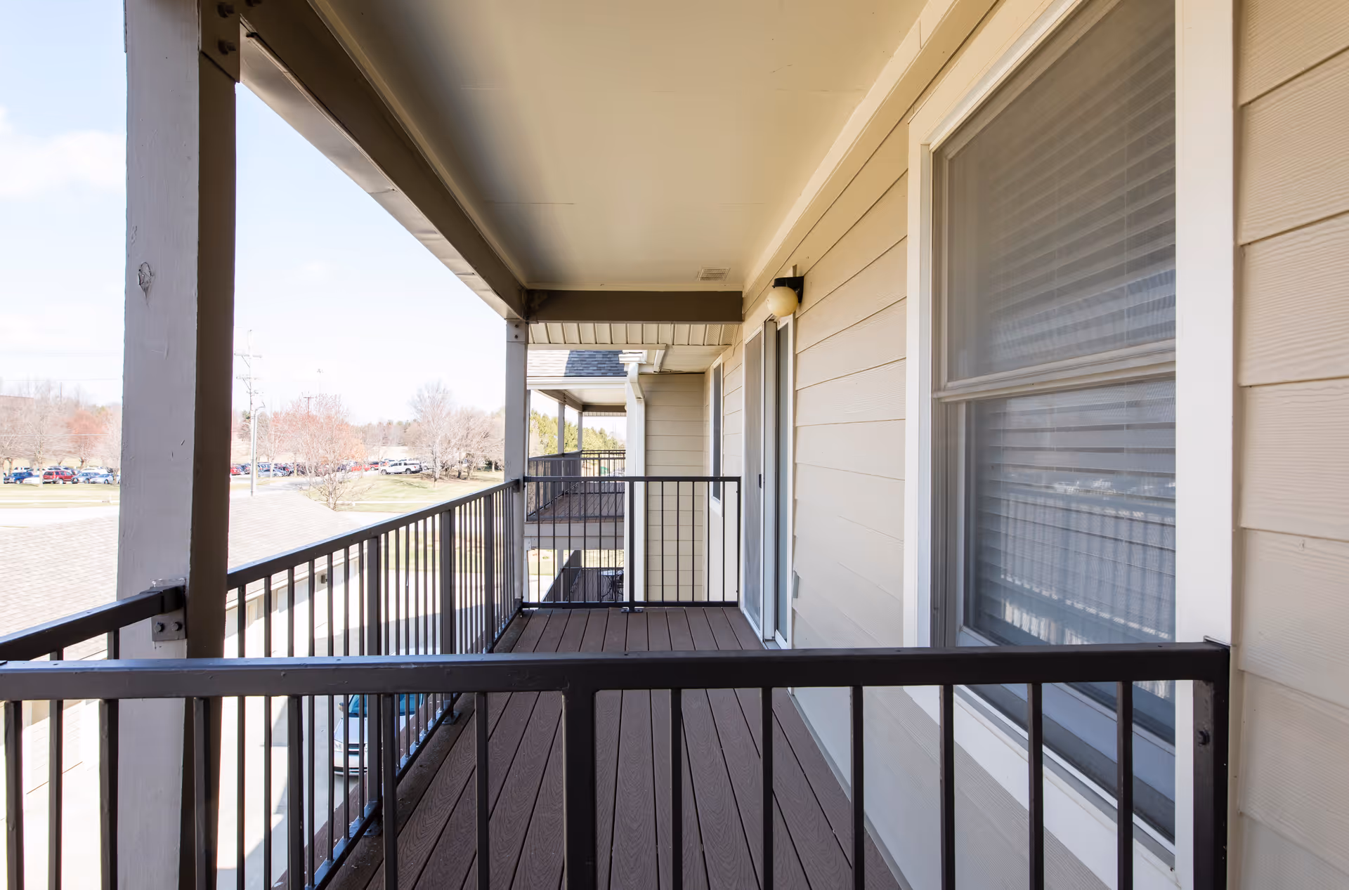 A covered balcony with brown wooden flooring and black metal railings. The balcony is attached to a beige building with a window and a sliding glass door. In the background, there is a view of trees, parked cars, and a clear sky.