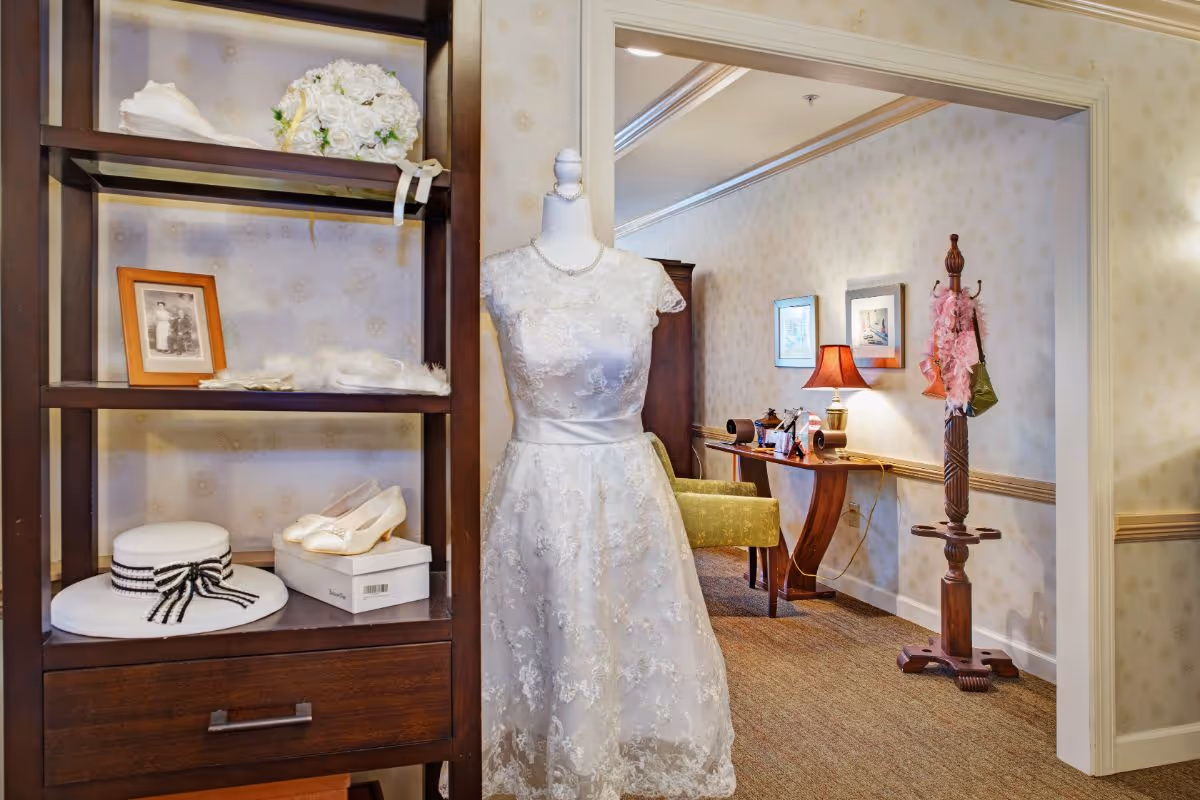 A mannequin in a white lace wedding dress stands beside shelving with a hat, shoes, and a framed photo in a warmly furnished interior room.