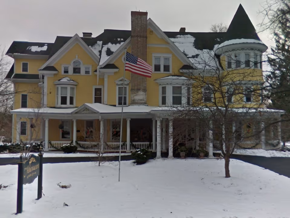 Yellow Victorian-style multi-story building with a wraparound porch, an American flag in front, and a snow-covered lawn.