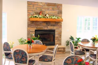 A cozy interior room with round wooden tables surrounded by floral upholstered chairs. A stone fireplace with a wooden mantel decorated with greenery and a clock is centered on the wall. Large windows allow natural light to brighten the space, and potted plants add a touch of greenery.