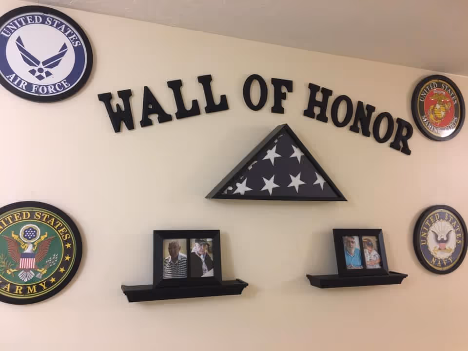A wall display titled 'WALL OF HONOR' featuring a folded American flag in a triangular case at the center. Surrounding the flag are four circular emblems representing the United States Air Force, Marine Corps, Army, and Navy. Below the flag are two small black shelves holding framed photos of elderly individuals.
