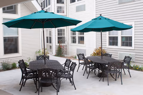 Outdoor patio area with two black metal tables, each surrounded by six matching chairs. Each table has a large green umbrella providing shade. The patio is adjacent to a light-colored building with multiple windows and some small plants along the edges.