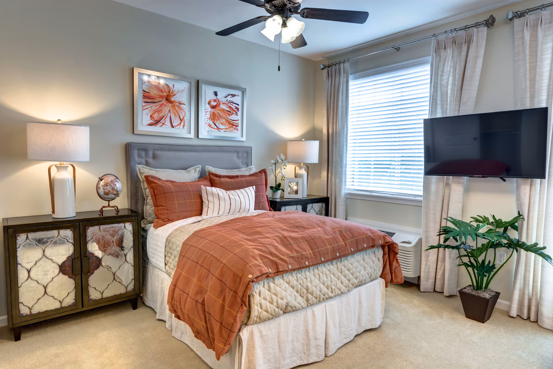 A cozy bedroom with a neatly made bed featuring rust-colored and beige bedding. Two framed floral artworks hang above the upholstered headboard. There are two bedside tables with lamps, a small globe, and a framed photo. A large window with white blinds and beige curtains lets in natural light. A flat-screen TV is mounted on the wall next to a green potted plant on the floor.