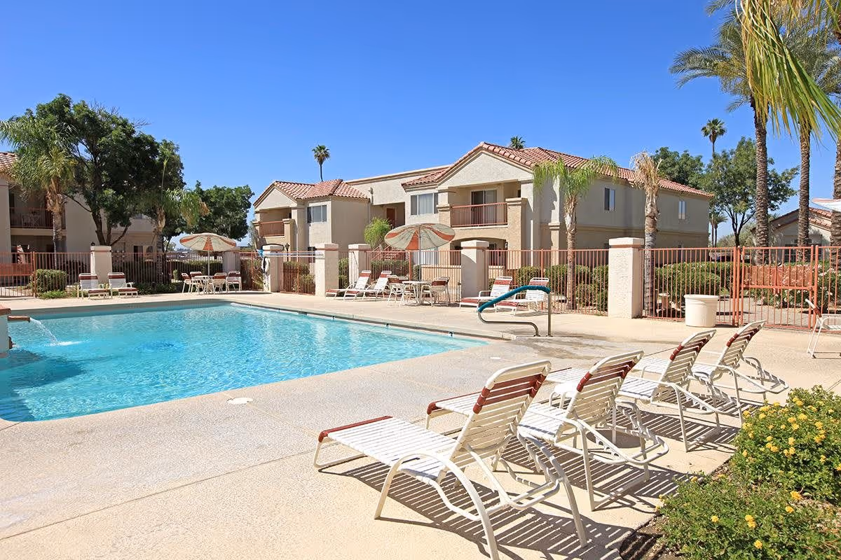 Outdoor swimming pool area at a senior living facility with several white lounge chairs lined up along the poolside. There are umbrellas and tables with chairs near the pool, surrounded by a fence. In the background, there are two-story beige buildings with red-tiled roofs and palm trees under a clear blue sky.