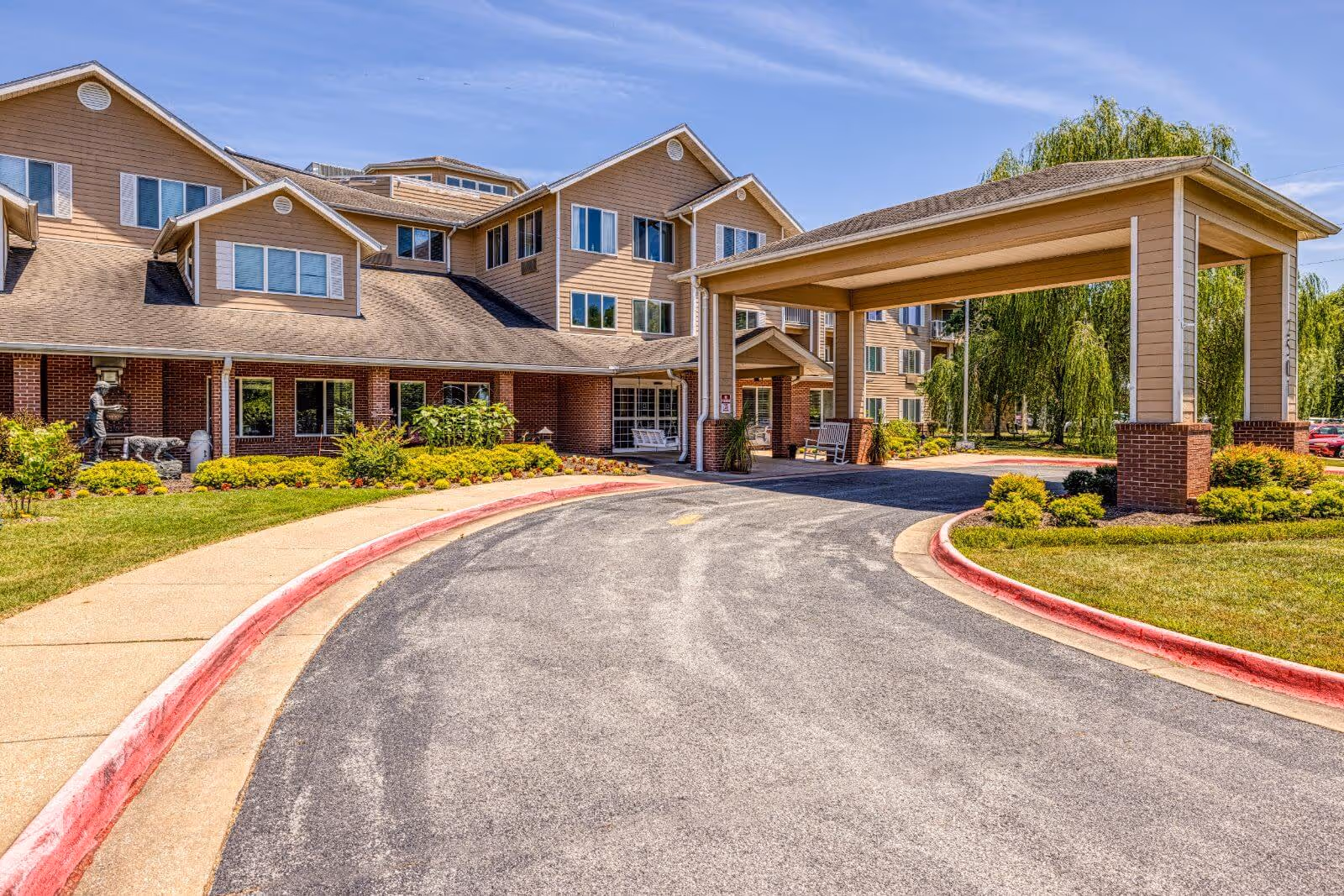 Exterior view of Morada Rogers senior living facility showing a multi-story building with beige siding and brick accents, a covered entrance with rocking chairs, landscaped bushes, and a curved driveway with red curbs under a clear blue sky.