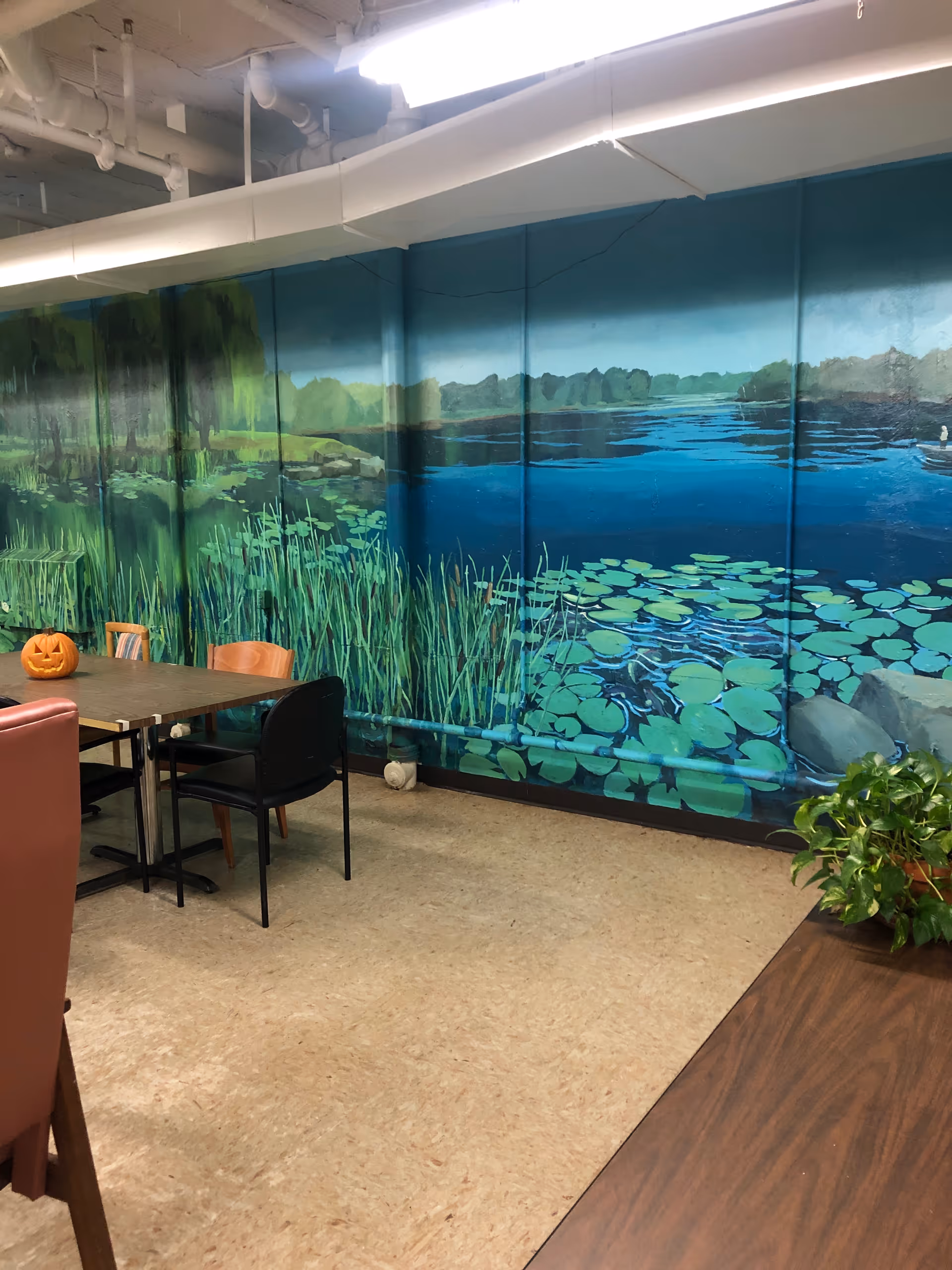 Indoor room with a mural painted on the wall depicting a pond with lily pads, tall grass, and trees in the background. There are several chairs around a table, one chair is black and another is wooden. A carved pumpkin is placed on the table. A potted plant is visible on a wooden surface in the foreground.