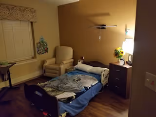 A small, cozy room with a single hospital-style bed covered with a blanket and some clothes. Next to the bed is a dark wooden nightstand with a lamp, a vase with sunflowers, and some personal items. There is a beige armchair near the window with closed blinds and a decorative valance. The walls are painted a warm beige color, and the floor is wooden.